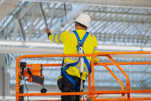 Certified construction worker wearing safety harness and high visibility vest operating an aerial work platform for elevated warehouse interior installation tasks