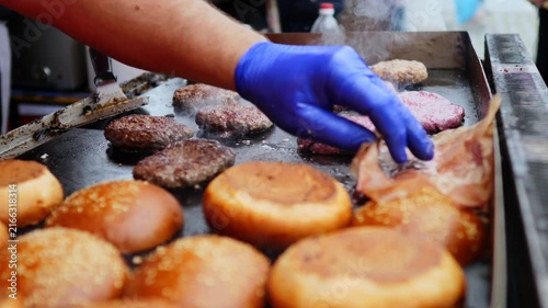 Street food chef wearing blue gloves preparing delicious burgers on a large, steaming flat top grill, cooking beef patties and toasting sesame buns for customers at a busy outdoor market