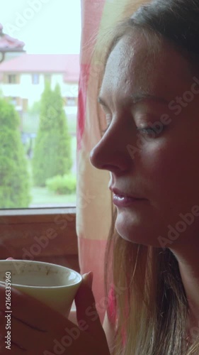 Sad female sitting by the window at home, holds mug with hot beverage. Vertical video