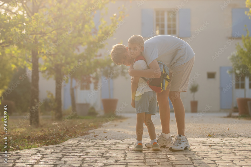 Fototapeta premium Father and son hugging, in outdoor cobblestone path residential area, with trees, house with blue shutters, cobblestone path, from a eye-level perspective perspective.