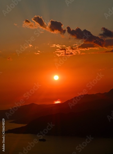 Canvas Print A vertical photograph featuring a bright glowing sun descending over dark mountain silhouettes and a calm sea bay, with dramatic clouds in the warm evening sky