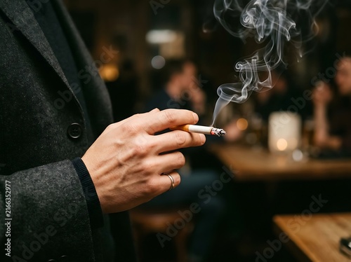 Man's hand holding burning cigarette with glowing embers and smoke rising against a dark warm backdrop.
