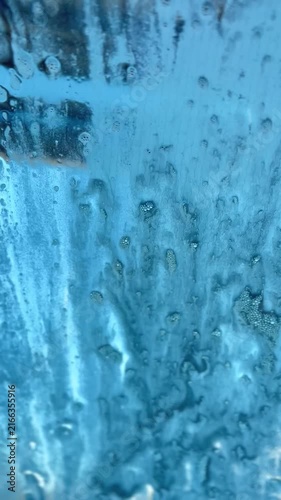 Thick blue soap foam and water droplets slide down a car windshield during an automated car wash cycle, creating an abstract liquid texture.