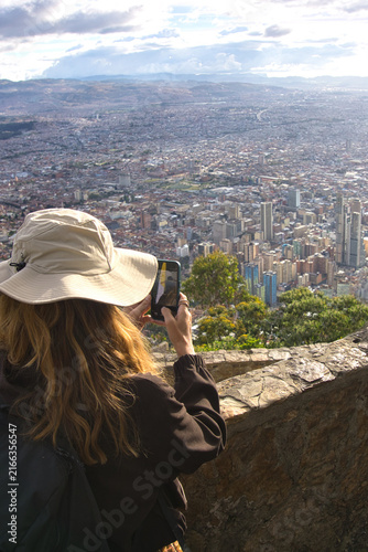 Fotografía Rear view of woman holding smartphone to photograph Bogota city skyline from mountain viewpoint at sunset