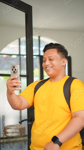 Young Asian man in a yellow shirt making a video call on his smartphone, smiling and waving his hand near a glass door indoors. Casual lifestyle and online communication concept, vertical format.