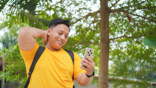 Happy young Asian man in a yellow shirt smiling shyly while looking at his smartphone, with one hand behind his neck outdoors. Casual lifestyle concept of receiving good news or a message.