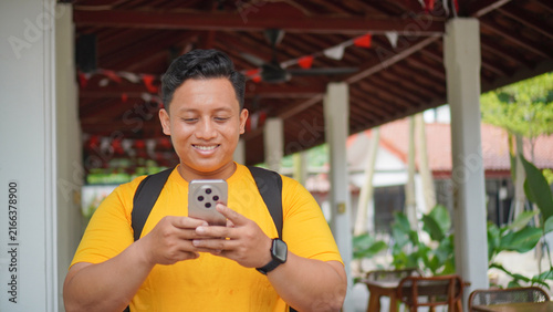Happy young Asian man in a yellow shirt smiling while looking at his smartphone, wearing a backpack and smartwatch under a wooden pavilion outdoors. Casual lifestyle and mobile technology concept.
