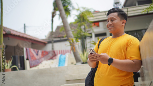 Happy young Asian man in a yellow shirt smiling while using his smartphone outdoors, wearing a backpack and smartwatch. Casual lifestyle concept of a student or traveler with copy space for text.