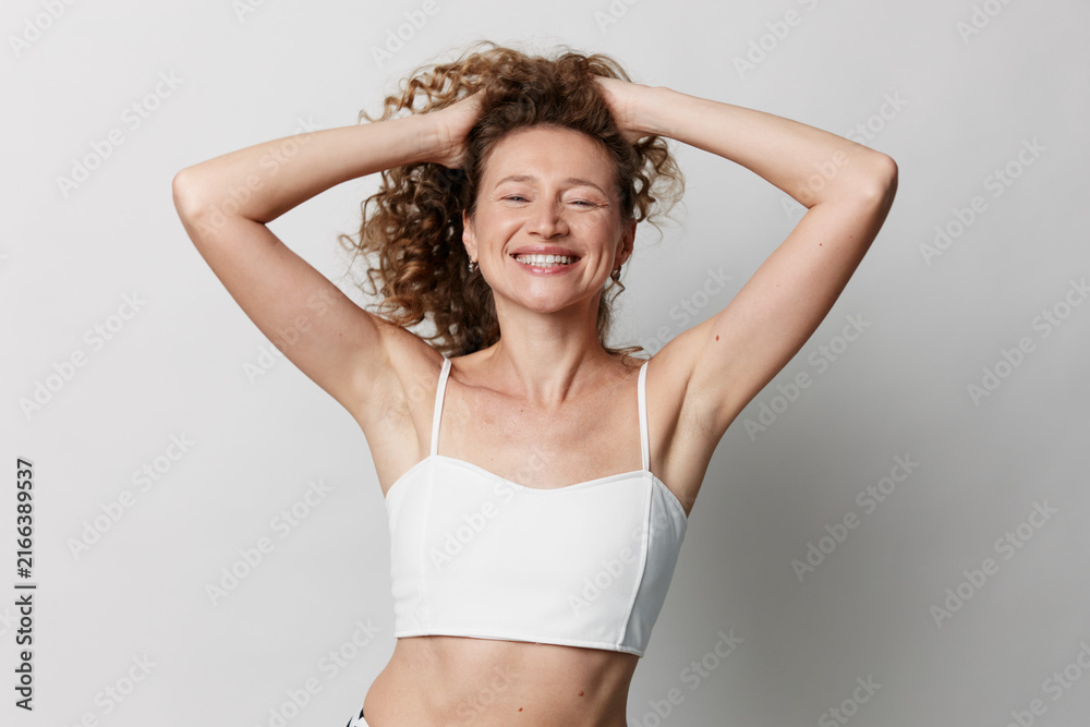 Fototapeta premium Authentic middle aged woman with natural skin smiles broadly while posing in a white crop top, arms raised, studio lighting, candid portrait.