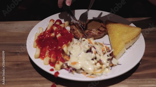 Person cutting grilled pork steak with knife and fork on white plate. Served with french fries with ketchup, fresh coleslaw, and toasted bread on wooden table. Delicious restaurant meal setup.