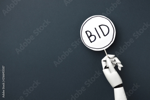 Konstfotografi A white robotic hand holding bidding paddle with BID signs on a a dark gray background