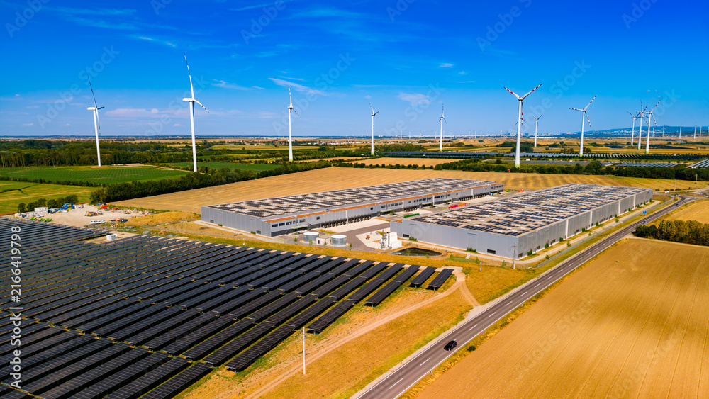 Fototapeta premium Renewable energy sources power a large facility in a rural area. Solar panels and wind turbines are visible beside a large building in an open field. The setting is bright and clear.