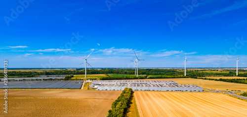 Wind turbines and solar panels in a rural landscape during the day. Wind turbines are turning above fields of crops and solar panels. A clear sky shows no clouds.