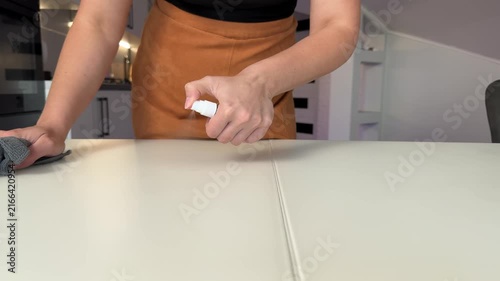Adult cleaning pale tabletop in modern kitchen, left hand pressing grey microfiber cloth along stitched seam while right hand holds small white spray bottle, with cleaning, wipe, spray, bottle detail