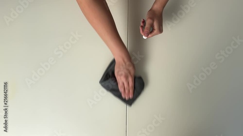 Hands cleaning seam between pale cream cabinet doors with microfiber cloth and clear spray bottle, neat household maintenance scene with soft neutral light and centered composition