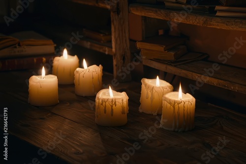 A warm still life of six lit pillar candles arranged on a rustic wooden table beside stacked books and wooden shelving. Soft flickering flames cast a golden