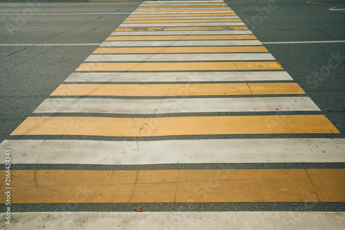 Close-up view of pedestrian crossing with alternating yellow and white painted stripes on dark asphalt, seen from low top-down angle with visible wear, scuffs, cracks, and weathered road texture