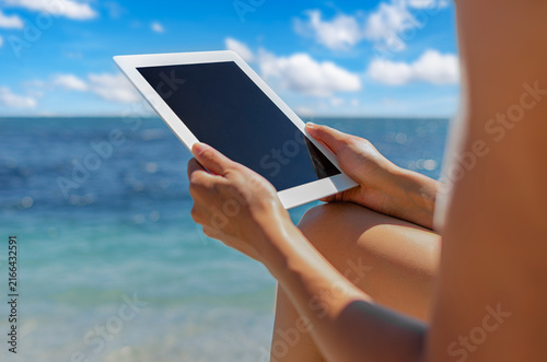 A person holds a digital tablet with a blank screen on a tropical beach.