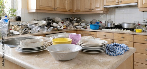 Plakat w ramie A messy kitchen countertop filled with dirty dishes and scattered kitchen tools in a dimly lit space.