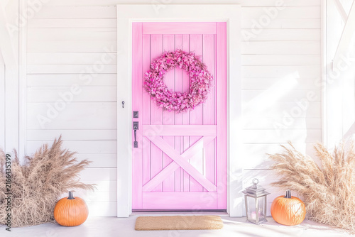 Cozy pastel pink front door decorated with floral wreath and pumpkins on porch background.