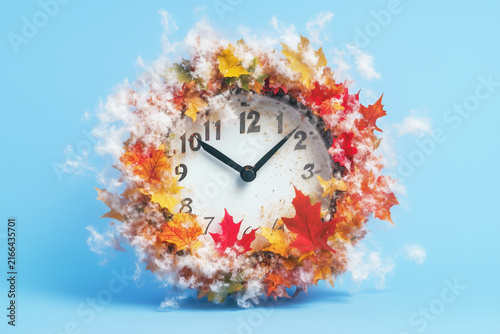 White wall clock surrounded by colorful autumn leaves and white fluff on a light blue background.