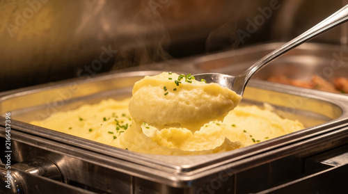 Close up of silver serving spoon scooping hot mashed potato from stainless steel buffet dish