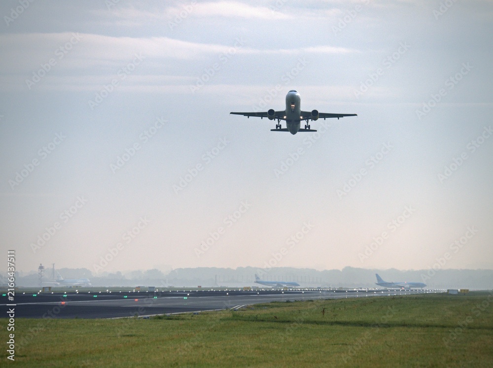 Fototapeta premium Airplane taking off from airport runway at sunrise with distant planes parked on the tarmac