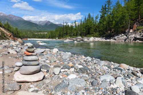Summer travel. Tourists built stone pyramid on bank of Irkut River on August day. Scenic landscape with river and Eastern Sayan Mountains in distance. Siberia, Buryatia, Tunka Valley, Nature Park