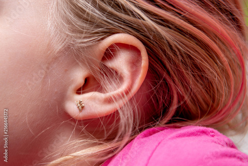 Close-up of a young child's ear wearing a delicate gold star stud earring, with pink hair and clothing, highlighting children's jewelry, first earrings, and childhood style.