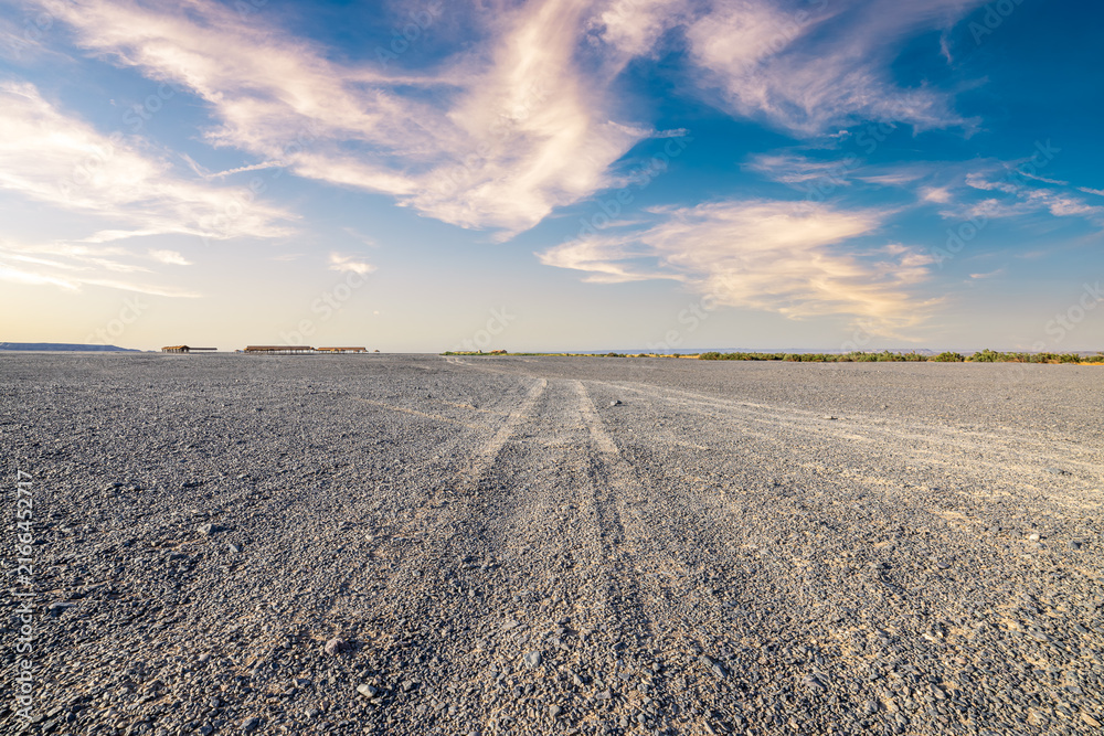 Naklejka premium Desert sand track leading through a vast gravel plain under a blue sky with wispy clouds in Xinjiang, China