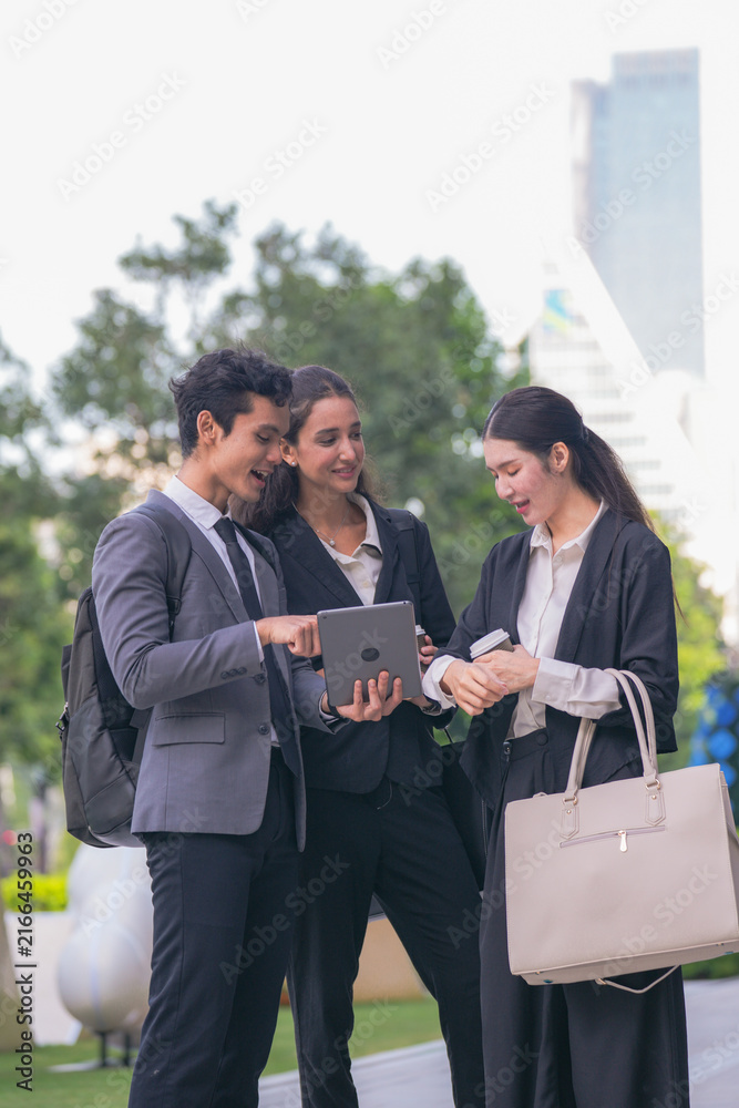 Fototapeta premium A diverse team of three business professionals comparing notes over a tablet screen in a green terrace with towers in the background, a modern picture of teamwork in a green city district