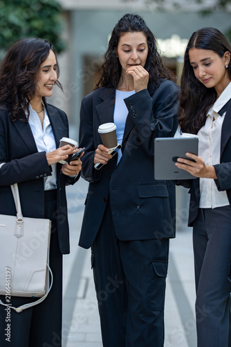 A team of three Hispanic businesswomen in matching dark suits, absorbed in the numbers on screen, hold an impromptu walking meeting over a tablet with a tablet screen shared between them.