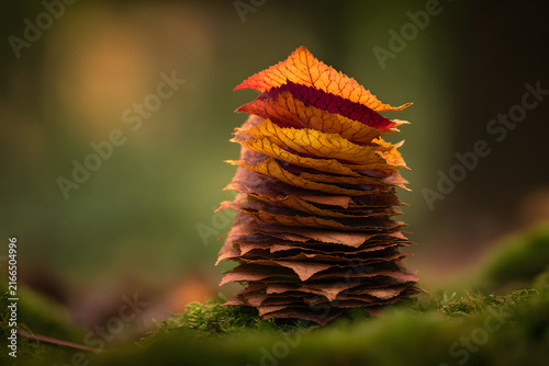 Close-up of a perfectly stacked pile of colorful autumn leaves, transitioning from bright orange and red to muted brown, set on vibrant green moss in a blurry woodland.