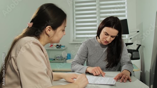 Woman doctor with patient fill medical records form together in clinic. Physician surveys brunette female to diagnose disease in hospital