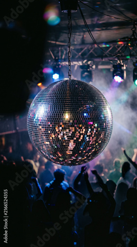 Ultra realistic close-up of a mirrored disco ball reflecting a crowded dance floor, silhouettes of young adults dancing under colorful beams of light 