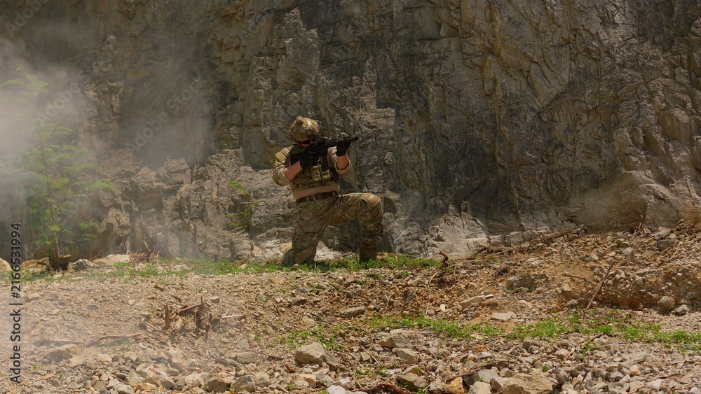 Obraz premium A special forces soldier in combat gear kneels with a rifle at the base of a cliff, maintaining vigilance during a tactical operation in a rugged environment.