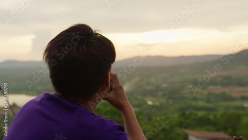 Konstfotografi A young man contemplates the future, enjoying a peaceful moment with a drink while overlooking a vast mountain range and valley during a beautiful sunset