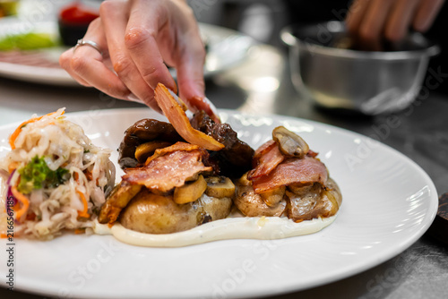 Obraz na plátne A hand places a piece of bacon onto a plate featuring roasted potatoes, mushrooms, beef, cream sauce, and a side salad