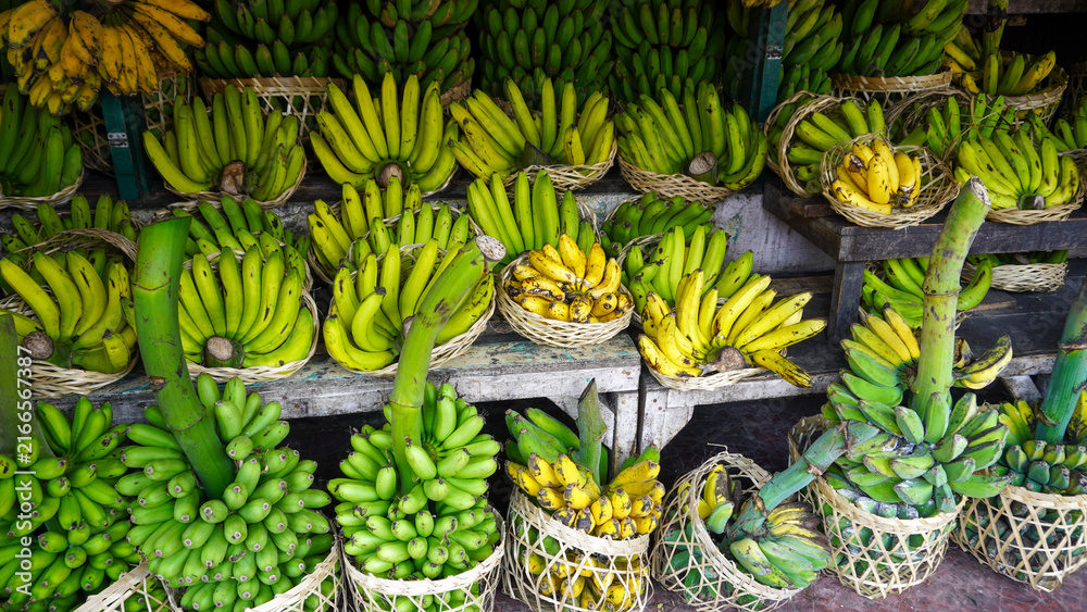 Fototapeta premium Fresh ripe and green bananas arranged in woven bamboo baskets at a traditional Asian fruit market stand. Organic tropical harvest stacked neatly on wooden shelves for local street market sale.
