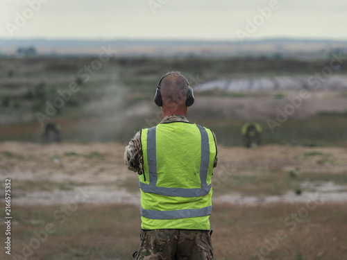 фотографія A range safety officer in a high visibility vest and ear defenders supervises live firing from behind the firing point on a military range