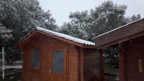 Snow falling around a wooden shed with snow covered roof and trees during cold overcast winter weather