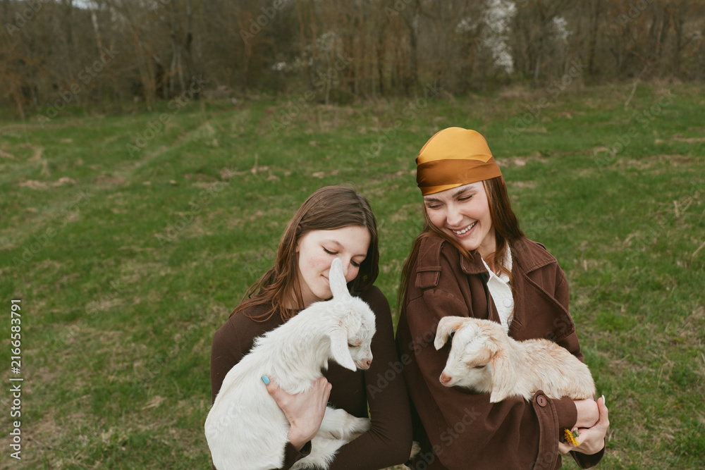 Fototapeta premium Two young women dressed in brown coats smile happily while holding white and light brown baby goats in a green meadow during a relaxed lifestyle moment outdoors.