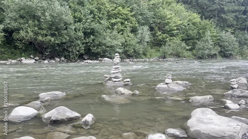 Calm mountain river flows around stacked balancing stones and smooth rocks near a green forest, creating peaceful travel, nature and meditation footage.