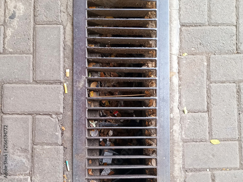Urban street drainage grate accumulating litter and cigarette butts, highlighting pollution and environmental neglect
