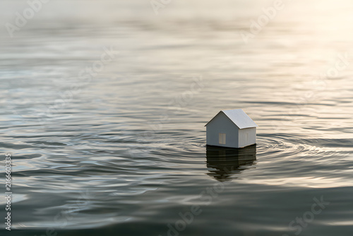 A simple white miniature house floats on calm blue water, reflecting its image. Represents themes of housing, displacement, or tranquility.
