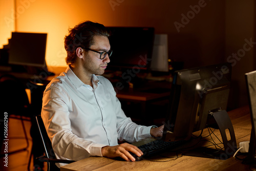 Stressed businessman rubbing eyes at desk in dim office after long workday, holding glasses with tired expression and warm blurred background during late evening