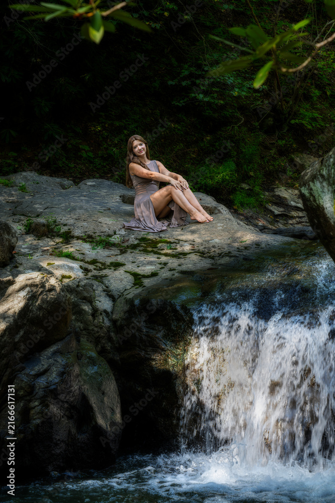 Fototapeta premium Young Woman Sitting on Mossy Rock Beside Waterfall in Lush Forest Stream, Relaxing Outdoors