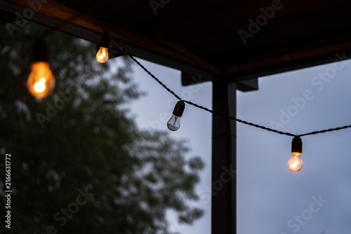 Quadro su tela Outdoor string lights with one faulty bulb hanging among illuminated lamps beneath a veranda roof, highlighting electrical maintenance, energy efficiency and troubleshooting concepts against