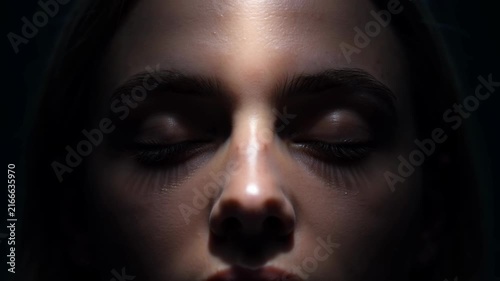 Close-up of a young woman's face with eyes closed and dramatic lighting casting shadows on her features, suggesting meditation, contemplation, or rest.