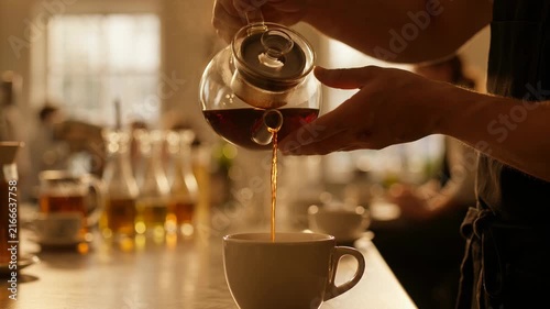 Barista pouring hot tea from a glass teapot into a white cup at a cafe counter, close-up shot in warm lighting